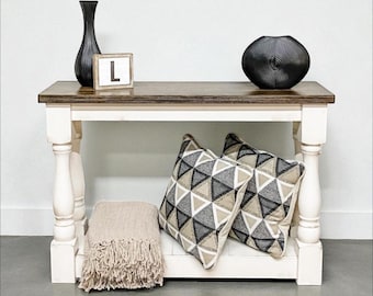 May include: A white and brown console table with decorative items. The table has a dark brown top and white base with turned legs. Two patterned pillows, a beige throw, and two black decorative vases are displayed on the table.