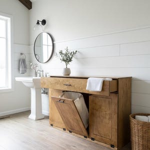 May include: A wooden bathroom cabinet with a laundry hamper, open drawer, and a basket. The cabinet is light brown with black hardware. A white towel is in the open drawer. The bathroom has a white sink, round mirror, and white shiplap walls.