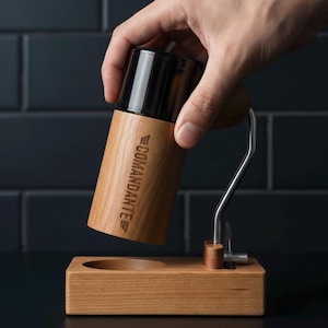 Peut inclure: Un moulin à café en bois avec un dessus noir et le mot "Comandante" gravé sur le côté. Le moulin est tenu au-dessus d'une base en bois avec une indentation circulaire. Une poignée en acier inoxydable est attachée au moulin.