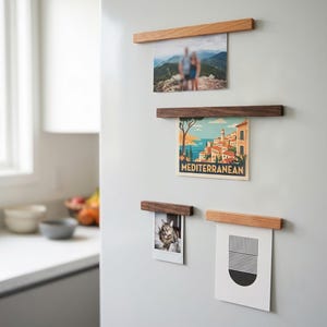 Könnte beinhalten: Vier rechteckige magnetische Holzleisten an einer flachen Oberfläche, die jeweils ein anderes Objekt halten: ein unscharfes Foto, eine bunte Mittelmeer-Reisepostkarte, ein kleines Katzenfoto und einen schwarz-weißen geometrischen Druck.