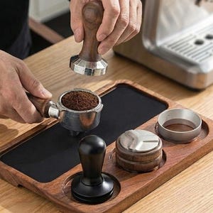 Op de afbeelding: Houten espressostation met een zwarte mat, een filterdrager gevuld met gemalen koffie en een stamper. Een persoon houdt de stamper boven de filterdrager. Andere accessoires zijn een doseerring en een distributietool.