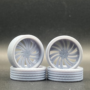 May include: Four light gray model car wheels. Each wheel has a tire with a tread pattern and a central hub with a spiral spoke design. The wheels are stacked in pairs, with two wheels on the bottom and two on top.