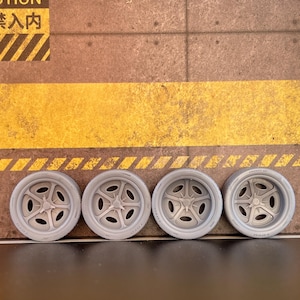 May include: Four gray toy car wheels with a five-spoke design are arranged in a row. The wheels are set against a textured, brown background with a yellow stripe and Japanese text.
