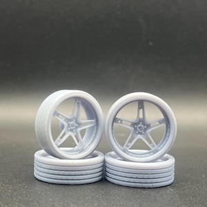 May include: Four light grey model car wheels and tyres are stacked on a dark surface. The wheels have a five-spoke design. The tyres have a tread pattern. The wheels and tyres are likely for a model car or a similar project.