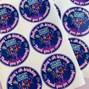 Puede incluir: Una hoja de pegatinas redondas con fondo azul oscuro. Cada pegatina tiene el texto "Audrey's 6th birthday" y "K-POP DEMON HUNTERS" en blanco y morado. Las pegatinas también presentan un gráfico de un grupo de personas y las palabras "Thank you for coming!"