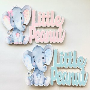 Puede incluir: Dos toppers de pastel de elefante gris con lunares rosas y azules. Los elefantes están sentados y tienen un lazo en la cabeza. El texto "Little Peanut" está escrito en purpurina rosa y azul.
