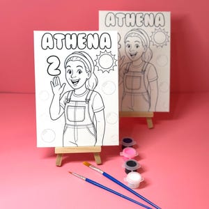 May include: Two white canvas boards with the name "ATHENA" and a cartoon illustration of a girl in overalls, with the number "2" on one. The boards are on small wooden easels. Paint pots and paintbrushes are arranged on a pink surface.