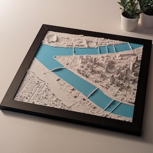 May include: A framed 3D map of a city, featuring white architectural models set against a blue background representing waterways. The map is encased in a black frame, with small potted plants in the background.