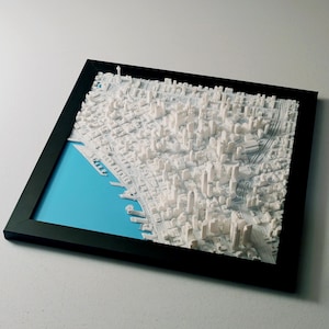 May include: A framed 3D relief map of a city, featuring white buildings and infrastructure against a blue background representing water. The map is encased in a black frame, creating a modern and architectural aesthetic.
