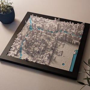 May include: A framed 3D relief map of a city, featuring white buildings and blue waterways. The map is set against a light blue background within a black frame. The detailed cityscape is a modern art piece.