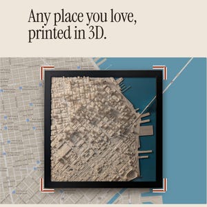 May include: A framed 3D printed city map model featuring a beige textured urban landscape with buildings and streets, set against a blue water section. The text "Any place you love, printed in 3D." is displayed above the black square frame.