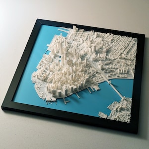 May include: A framed 3D relief map of a city, likely New York, with white buildings and infrastructure set against a blue background. The map is encased in a black frame, creating a striking contrast. The detailed model showcases the city's layout.