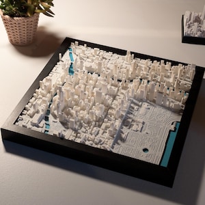 May include: A square framed 3D map of a city featuring white miniature buildings and structures set against a bright blue base. The detailed urban layout includes dense skyscraper blocks and a large park area, all enclosed within a black frame.