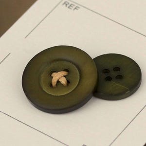 May include: Two olive green wooden buttons. One button has a tan thread detail in the center, and the other has four holes. The buttons are on a white surface with the word "REF" visible.