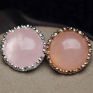 Pode incluir: Dois cabochões redondos de pedras preciosas, um com moldura prateada e outro com moldura dourada. As pedras preciosas são de cor rosa suave e as molduras têm uma borda decorativa e texturizada. O fundo é de um tom neutro e desfocado.