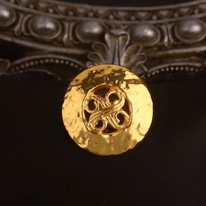 Könnte beinhalten: Ein goldfarbener, gehammerter Metallknopf mit einem komplizierten, wirbelnden Design in der Mitte. Der Knopf ist rund und hat eine strukturierte Oberfläche. Der Hintergrund ist schwarz, mit einem silberfarbenen Dekorationselement oben.