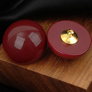 Puede incluir: Dos botones redondos de color rojo intenso con un acabado liso y brillante. Un botón muestra su superficie frontal sólida y abovedada, mientras que el otro está volteado para revelar su parte posterior plana con un enganche de metal dorado.