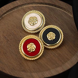 May include: Three round buttons with gold-colored rims and a rope-like texture. Each button has a different center color: cream, black, and red. A gold-colored rose is in the center of each button. The buttons are on a wooden surface.