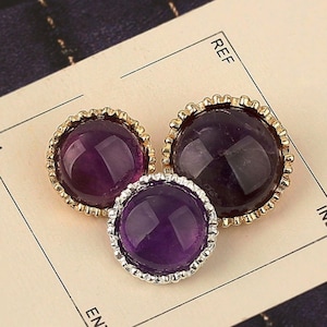 May include: Three round amethyst gemstones with gold and silver-colored decorative settings. The gemstones are a deep purple color and are set on a light beige surface. The settings have a beaded edge.