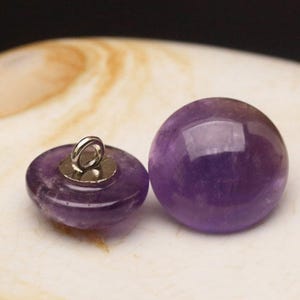 Könnte beinhalten: Zwei Amethyst-Edelsteinperlen. Eine hat eine ovale Form mit einer silbernen Schlaufe, die andere ist eine glatte Kugel. Die Edelsteine sind tiefviolett und haben eine polierte, reflektierende Oberfläche.