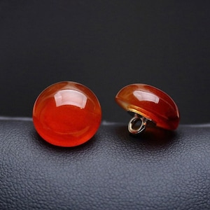 May include: Two glossy, orange-red buttons. One is fully round, the other a half-sphere with a metal loop. The buttons are set against a dark, textured background. The buttons are likely for clothing.