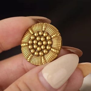 May include: A gold button with an ornate design. The button has a central cluster of small spheres surrounded by a textured, circular border. The button is held between fingers, highlighting its detailed craftsmanship.