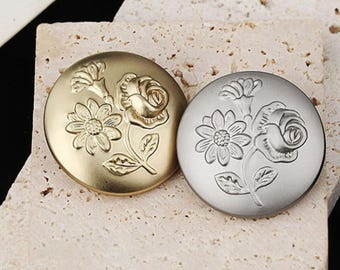 May include: Two decorative buttons with a floral design. The button on the left is gold and the button on the right is silver.