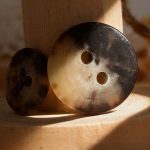 以下が含まれることがあります： 2つの大きな丸いボタンと、それぞれ2つの穴、そして1つの穴の小さなボタン。ボタンはマーブル模様で、薄茶色と濃い茶色の色が混ざっています。ボタンは天然素材で作られています。