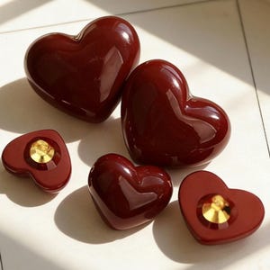 Puede incluir: Un conjunto de cinco botones en forma de corazón con un acabado en rojo burdeos brillante. Dos de los botones se muestran con sus enganches metálicos dorados hacia arriba, mientras que los otros tres muestran sus partes superiores redondeadas.