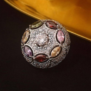 Op de afbeelding: Een zilverkleurige ring met een centrale heldere edelsteen omringd door kleinere heldere stenen. De ring heeft ovaalvormige, veelkleurige edelstenen in rood, roze, geel, groen en paars. Op een gouden en zwarte achtergrond.
