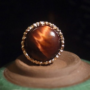 May include: A round, gold-toned ring featuring a large, polished, brown gemstone with amber and dark brown striations. The gemstone is set in a decorative, scalloped bezel. The ring is displayed on a brown surface against a dark background.