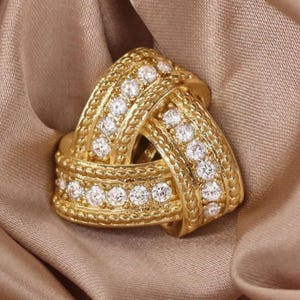 Op de afbeelding: Een gouden ring met een knoopdesign en een rij kleine diamanten rond de rand.