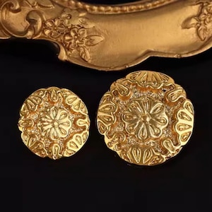 Puede incluir: Dos botones dorados y ornamentados con un diseño floral. Los botones son redondos y presentan patrones de relieve detallados. El fondo es negro, con un vistazo a un marco dorado decorativo en la parte superior.