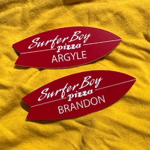 May include: Two red surfboard-shaped stickers with white text. The top sticker says "Surfer Boy Pizza Argyle" and the bottom sticker says "Surfer Boy Pizza Brandon".