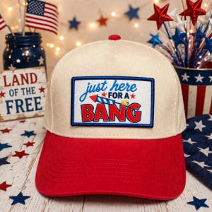 May include: A cream-colored baseball cap with a red brim and a rectangular patch on the front. The patch features the blue text "just here" above the red text "FOR A" and the large red text "BANG" with a firework graphic and small red stars.