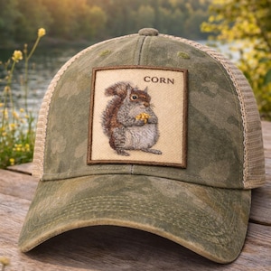 May include: A green camouflage baseball cap with a tan mesh back. The cap features a square patch with a detailed illustration of a squirrel holding corn, with the word "CORN" above the squirrel.