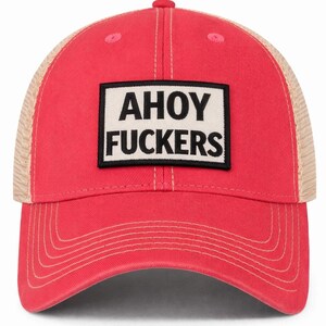 May include: A bright red baseball cap with a tan mesh back and a rectangular patch on the front. The patch is white with a black border and features the black text AHOY FUCKERS in a bold, sans-serif font. The cap has matching red stitching.