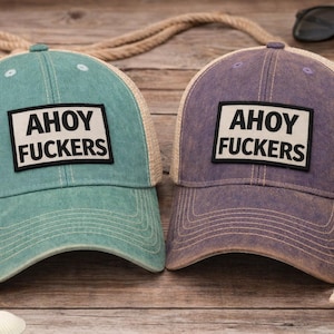 May include: Two baseball caps with mesh backs, one in teal and one in purple. Each cap features a rectangular patch on the front with the text AHOY FUCKERS in bold black letters. The caps have a distressed finish and a curved brim with stitching.