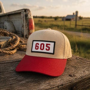 May include: A beige baseball cap with a red brim. The cap features a rectangular patch with the number "605" in red on a white background, framed by a black border. The hat is set on a wooden surface.