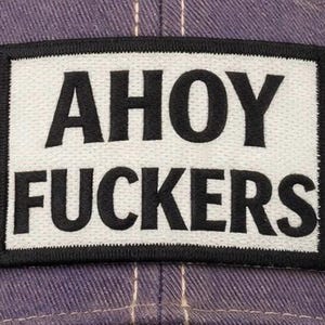 May include: A rectangular embroidered patch featuring a white background with a thick black border. The text AHOY FUCKERS is stitched in bold black capital letters across the center. The patch is attached to a purple fabric surface with visible stitching.