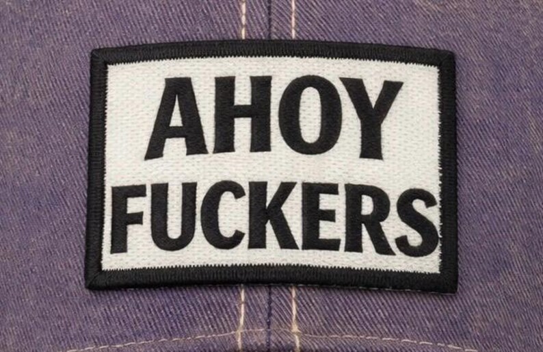 May include: A rectangular embroidered patch featuring a white background with a thick black border. The text AHOY FUCKERS is stitched in bold black capital letters across the center. The patch is attached to a purple fabric surface with visible stitching.