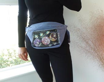 May include: A light blue fanny pack with a clear front pocket displaying several colorful pinback buttons. The fanny pack is worn around the waist and has a zippered closure. The buttons feature various designs and colors.