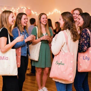 May include: A group of people are gathered, some holding drinks, with several carrying tote bags. The bags are either light pink or off-white, decorated with small white flowers. Some bags have names like "Mia", "Sophie", and "Lily" printed on them.