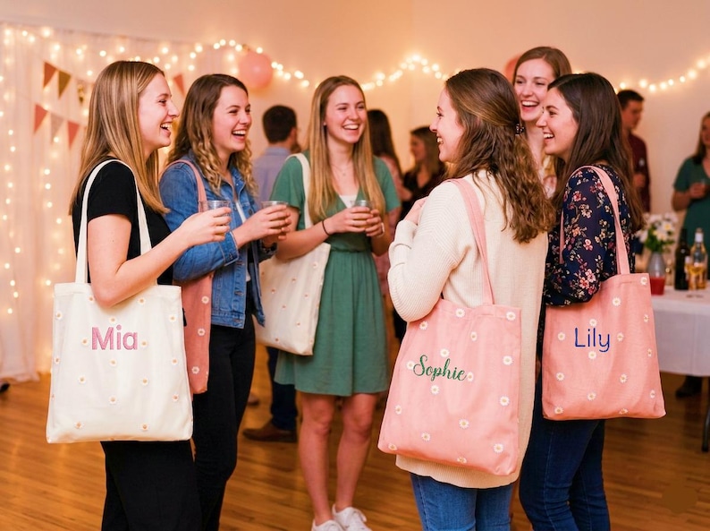 May include: A group of people are gathered, some holding drinks, with several carrying tote bags. The bags are either light pink or off-white, decorated with small white flowers. Some bags have names like "Mia", "Sophie", and "Lily" printed on them.