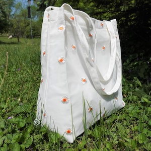 May include: White canvas tote bag with embroidered orange and white daisy flowers. The bag has two white handles and is sitting in a grassy field. The tote bag is a reusable shopping bag.