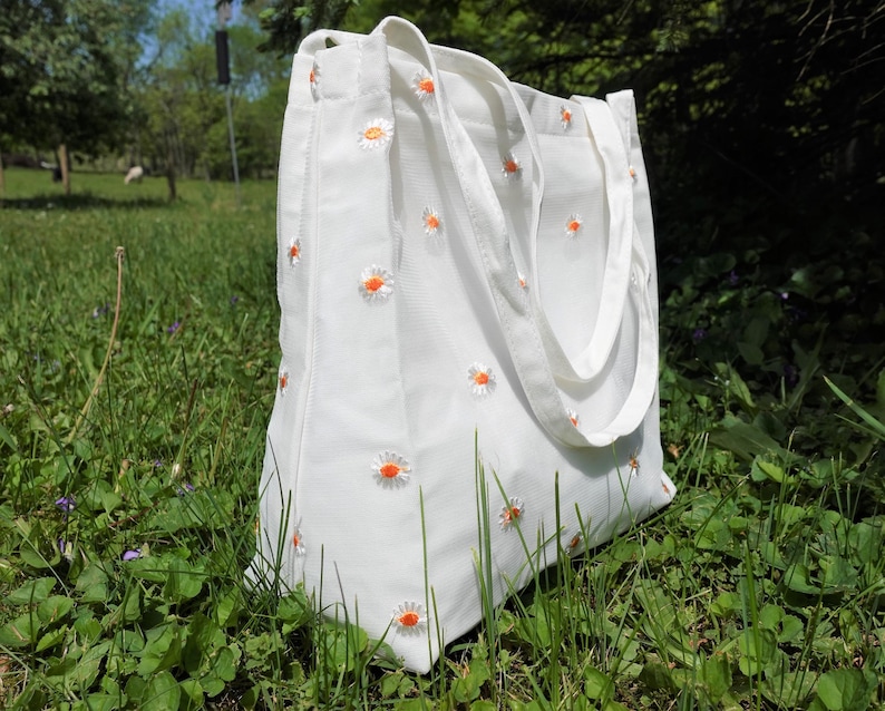 May include: White canvas tote bag with embroidered orange and white daisy flowers. The bag has two white handles and is sitting in a grassy field. The tote bag is a reusable shopping bag.