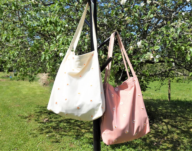 May include: Two canvas tote bags hanging outdoors. One is cream-colored with small orange daisy patterns, and the other is pink with white daisy patterns. Both bags have long straps and are suspended from a black metal pole, with a green lawn and tree in the background.