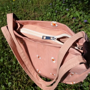 May include: A dusty rose-colored shoulder bag with a white zipper and daisy embroidery. The bag has a rectangular shape with a long strap and is open, revealing the interior lining. The bag is sitting on a bed of green grass.