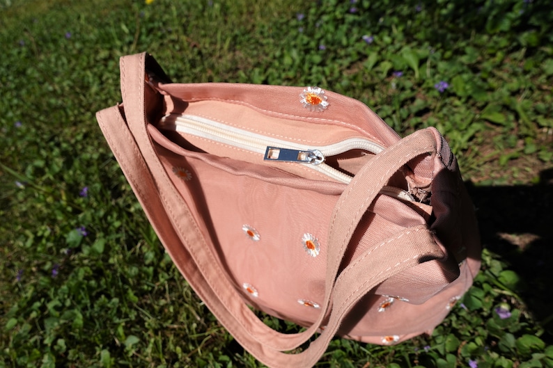 May include: A dusty rose-colored shoulder bag with a white zipper and daisy embroidery. The bag has a rectangular shape with a long strap and is open, revealing the interior lining. The bag is sitting on a bed of green grass.