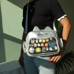 May include: Silver cat-shaped crossbody bag with a clear front pocket filled with colourful character pins. The bag has a silver strap and cat ear details. The person is wearing a black top and blue jeans.